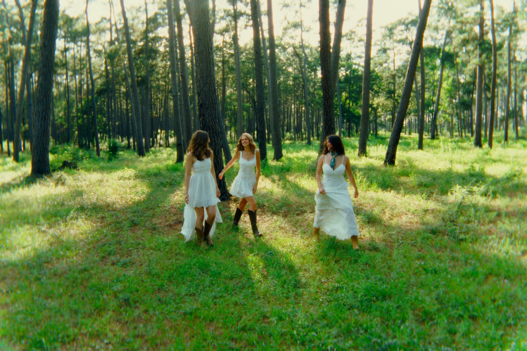 Three women in flowing white dresses walking through a sunlit forest